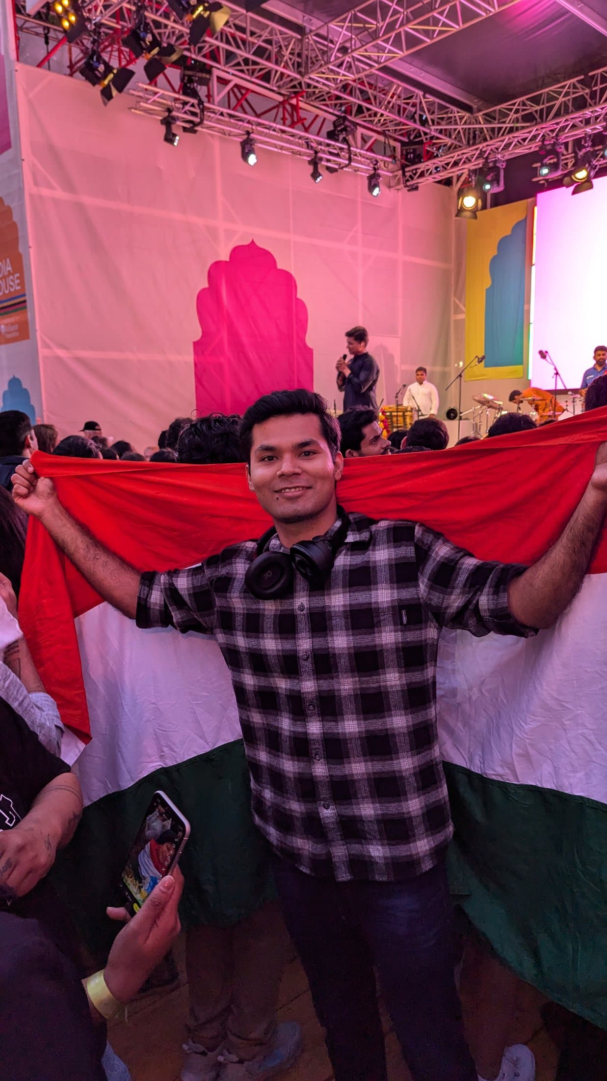Shivam smiling and holding up the Indian flag at a lively evening event during the Paris 2024 Olympics.