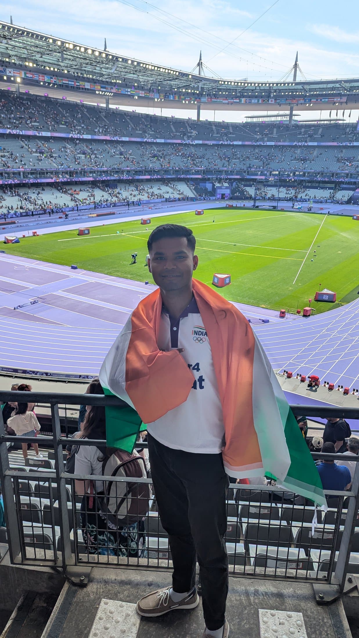 Shivam draped in the Indian flag inside an Olympic stadium, with the running track and pitch visible below.
