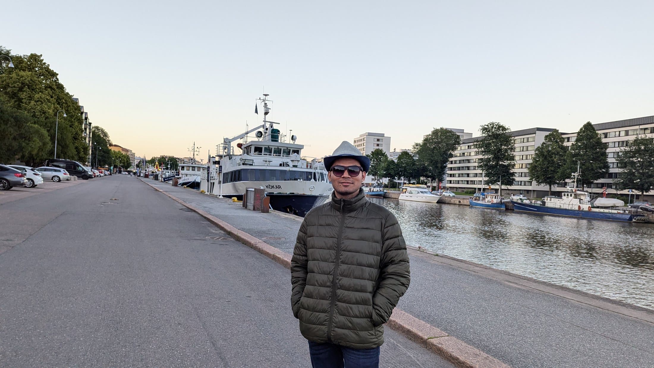 Shivam standing along a quiet Finnish harbor at dusk with a docked boat and waterfront buildings in soft evening light.