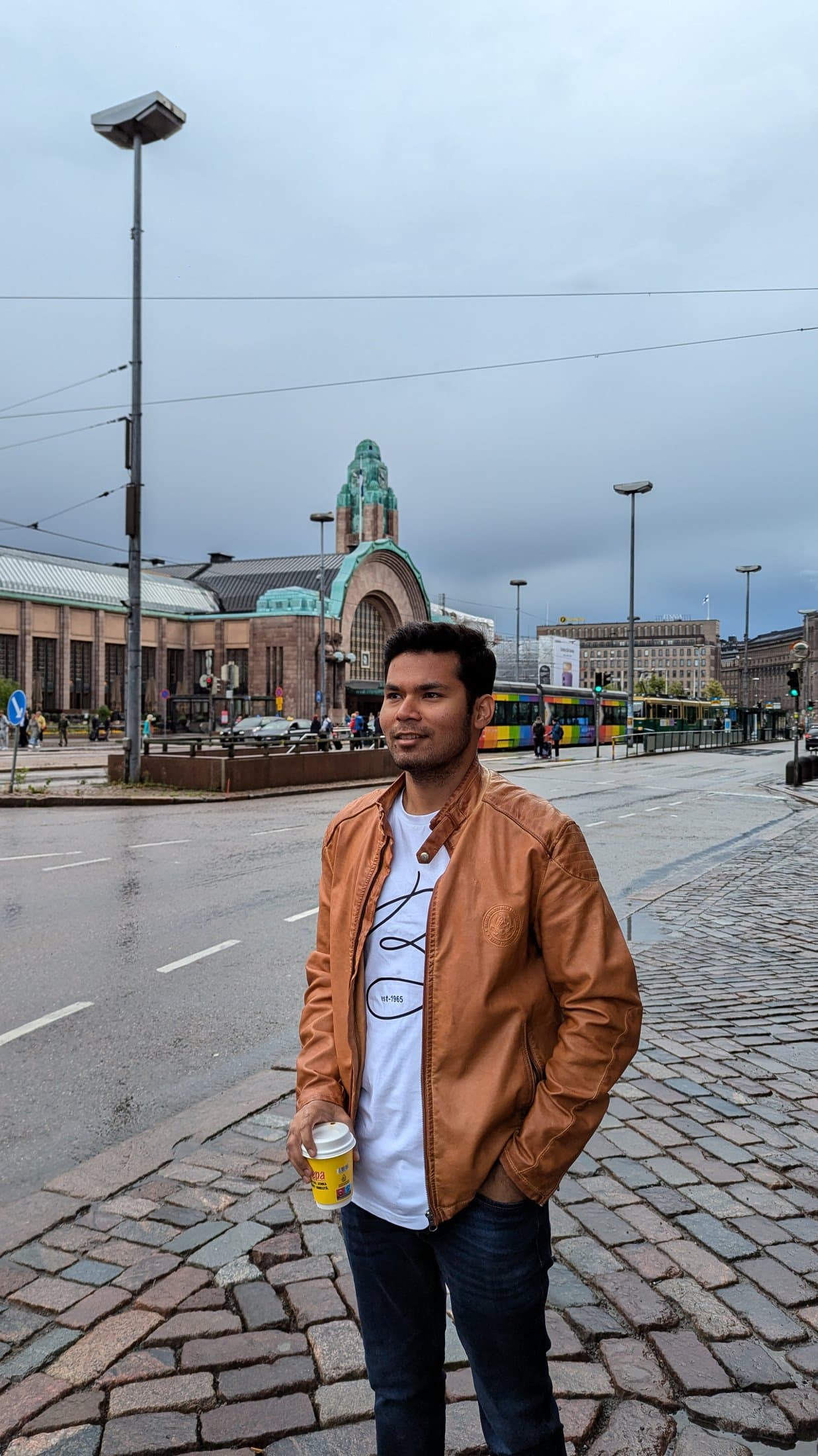 Shivam walking down a rain-slicked cobblestone street in Finland past a domed building, holding a drink.