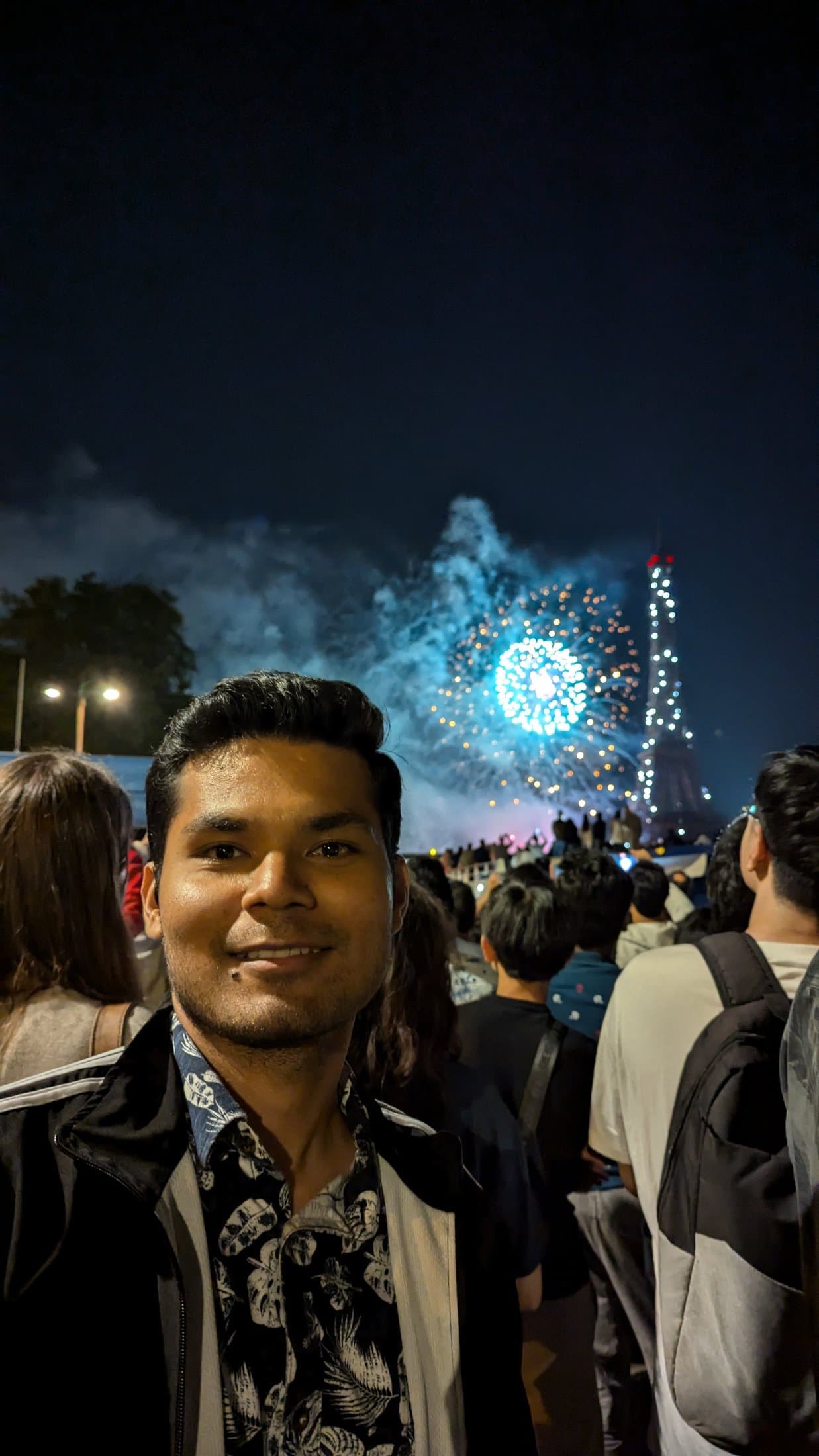 Shivam smiling at night in a crowd in Paris with fireworks lighting up the sky behind him.