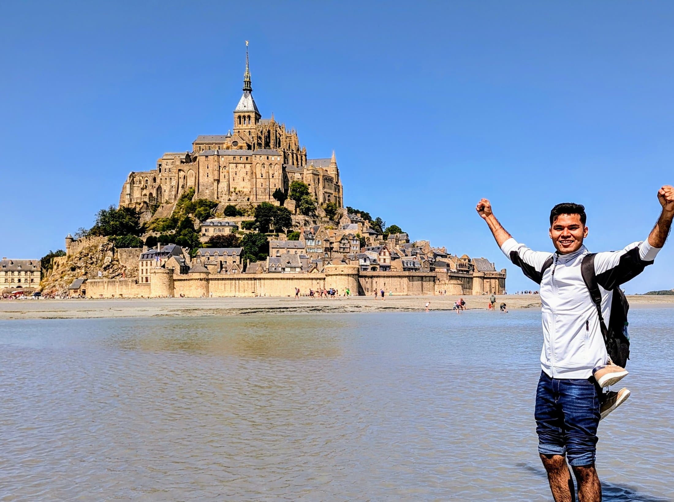Shivam with arms raised on the tidal flats facing Mont Saint-Michel, the fortified abbey rising from the water under a clear blue sky.