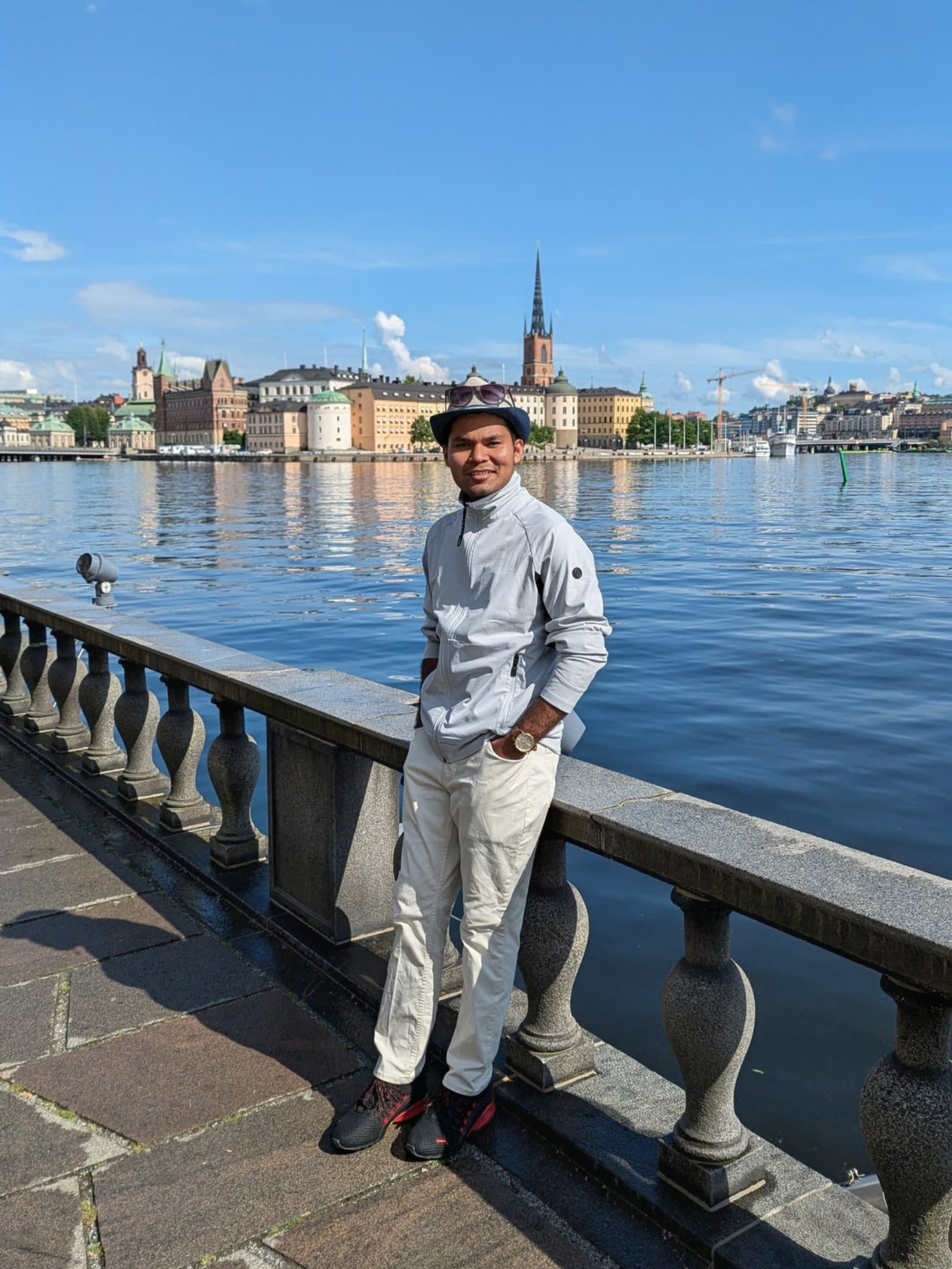 Shivam leaning on a stone railing along the Stockholm waterfront with the colorful buildings of Gamla Stan across the water.